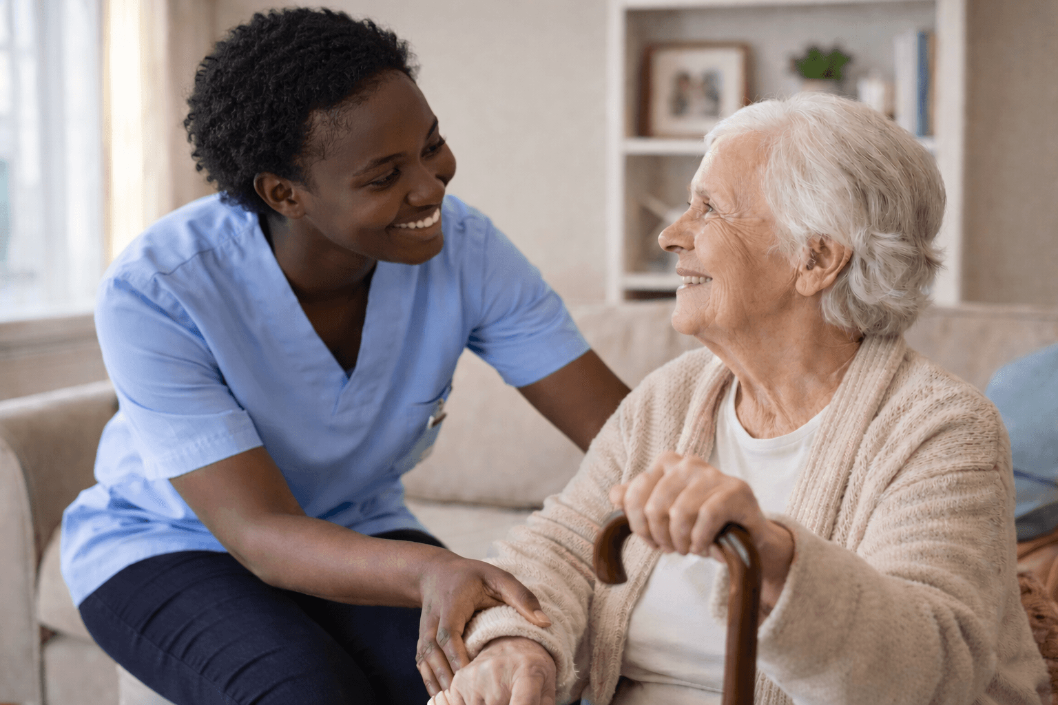 Smiling young Black caregiver in blue scrubs supports an elderly woman sitting with a cane.
